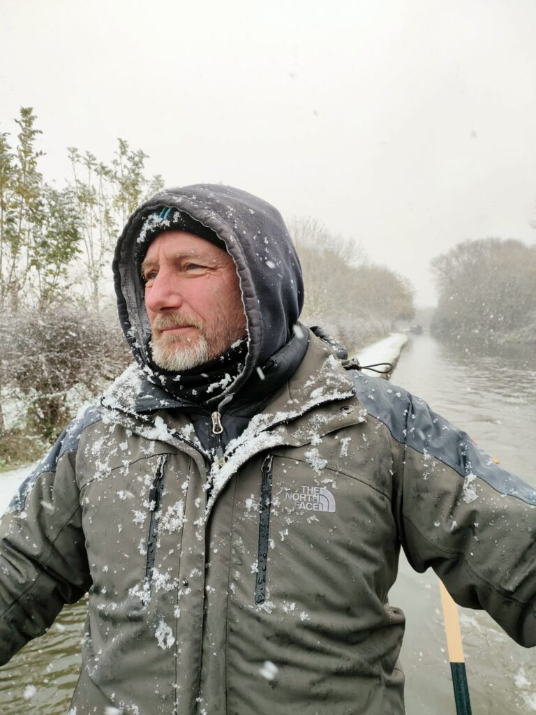 Image of Dr Steve Allen, A middle aged man driving a boat in a snow storm. He has snot on his jacket and in his beard. Behind him is the canal and snow covered trees